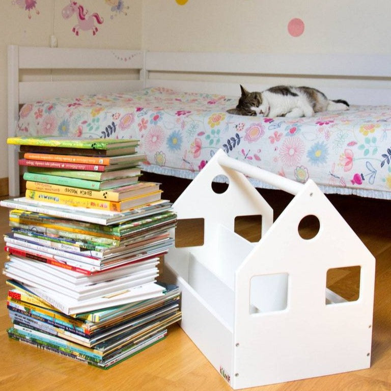 Empty white house-shaped book storage bin beside a stack of books.